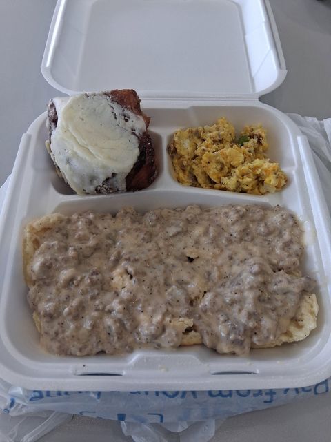 Takeout container with biscuits covered in sausage gravy, scrambled eggs and a frosted pastry